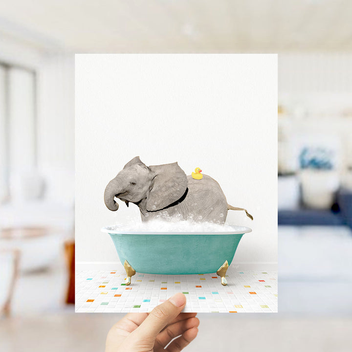 A hand holds up a framed image depicting a gray elephant sitting in a green bathtub, with a yellow rubber duck on its back.
