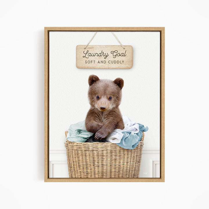 A cute brown bear sitting in a wicker laundry basket, with a wooden sign above that says "Laundry Goal Soft and Cuddly".