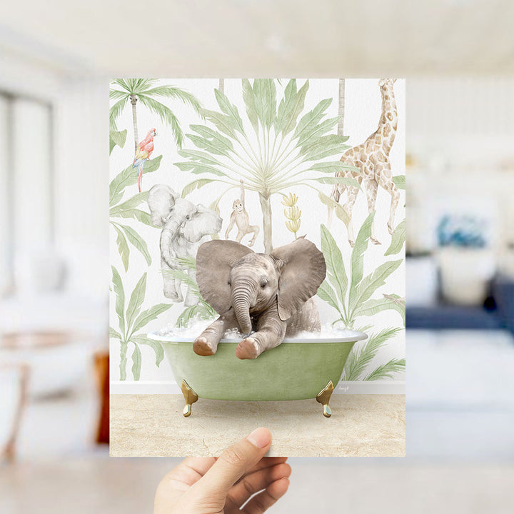 A hand holds up a framed print featuring an elephant sitting in a bathtub surrounded by tropical plants and animals, including a giraffe and a parrot.
