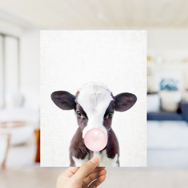 A hand is holding up a picture of a cow blowing a bubble with a pink bubble gum bubble.