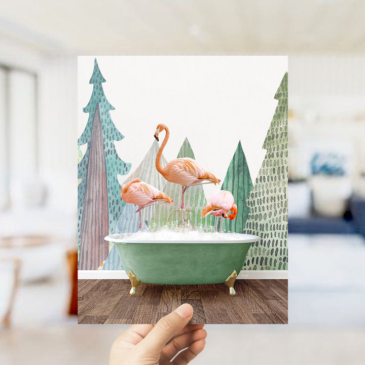 A hand holds up a framed image depicting three flamingos standing in a green bathtub, with a backdrop of trees and a snowy landscape.