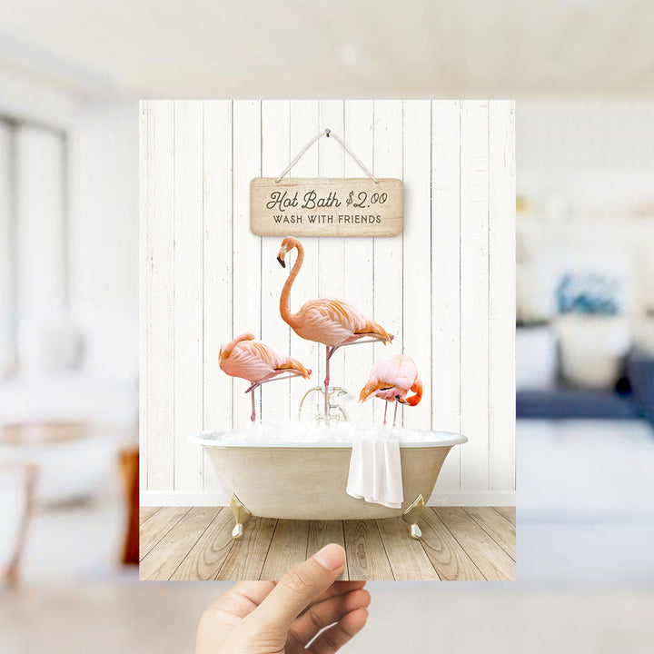 A hand holds up a framed image of a hot bath with three flamingos sitting in it, against a white wooden wall.