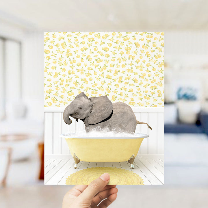 A hand holds up a framed image depicting a gray elephant sitting in a yellow bathtub filled with bubbles, surrounded by a yellow and white floral pattern.