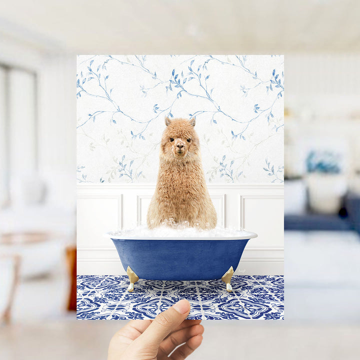 A hand holds up a framed image of a brown alpaca sitting in a blue bathtub filled with bubbles, set against a blue and white floral wallpaper.