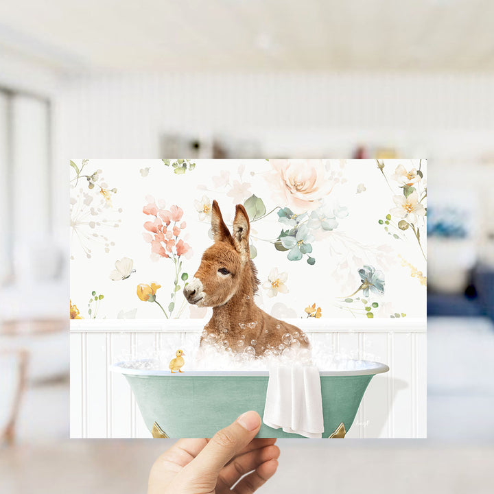 A hand holding a framed print featuring a brown rabbit sitting in a green bathtub filled with bubbles, surrounded by a floral pattern.