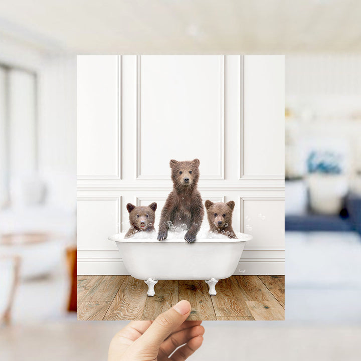 A hand holds up a framed photograph of three adorable brown bears sitting in a white bathtub, with the bears appearing to be enjoying a bath.