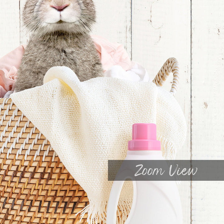 A cute gray rabbit sits in a woven basket filled with laundry, with a bottle of laundry detergent next to it.