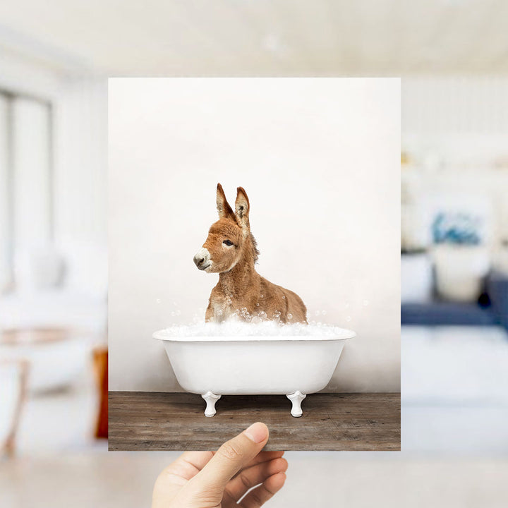 A hand holds up a framed image of a donkey sitting in a bathtub filled with bubbles.