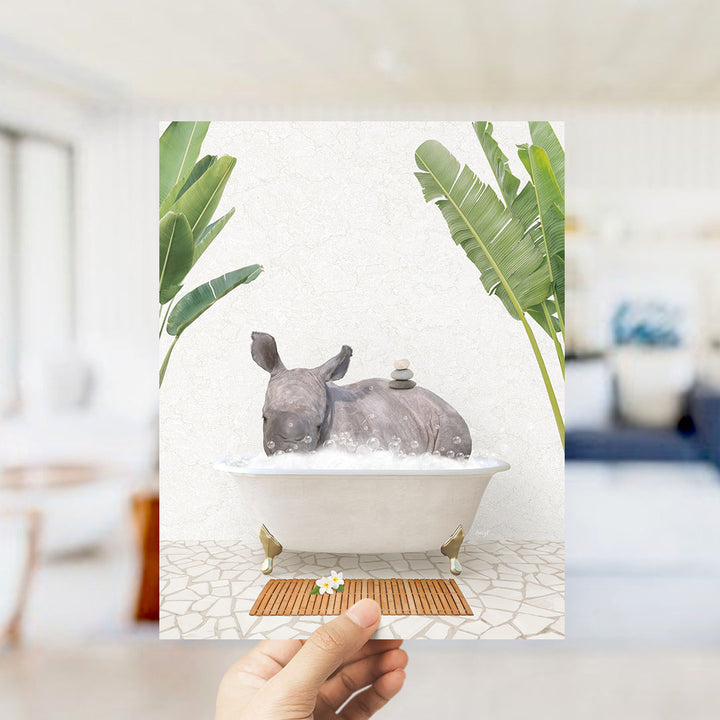 A hand holds up a framed image depicting a rhinoceros sitting in a bathtub filled with bubbles, surrounded by tropical plants.