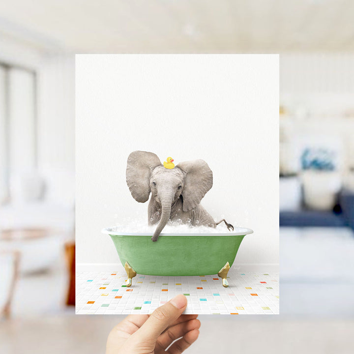 A hand holds up a framed image of a baby elephant sitting in a green bathtub filled with bubbles, wearing a yellow rubber duck.