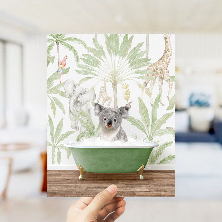 A hand holds up a framed image depicting a koala sitting in a green bathtub, surrounded by tropical foliage and animals.