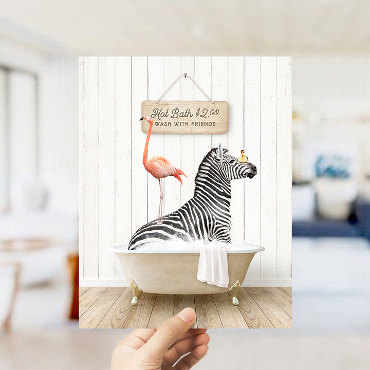 A hand holding a framed print featuring a zebra sitting in a bathtub, with a flamingo standing on its back and a sign above the bathtub that reads "Hot Bath $2.00 WASH WITH FRIENDS".