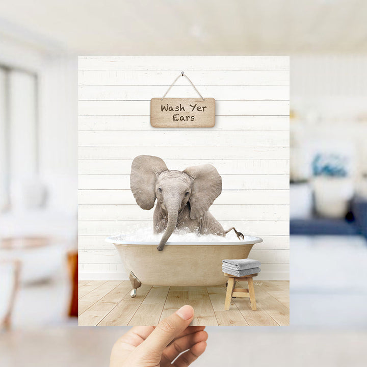 A hand holds up a framed image of an elephant sitting in a bathtub, with a sign above it that says "Wash Your Ears".