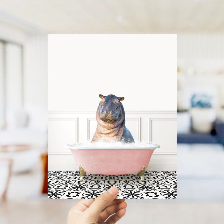 A hand holds up a framed image of a hippopotamus sitting in a pink bathtub filled with bubbles, surrounded by a black and white patterned floor.