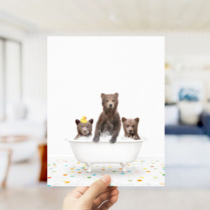 A hand holds up a framed image depicting three baby bears sitting in a bathtub, with one bear wearing a yellow rubber duck.
