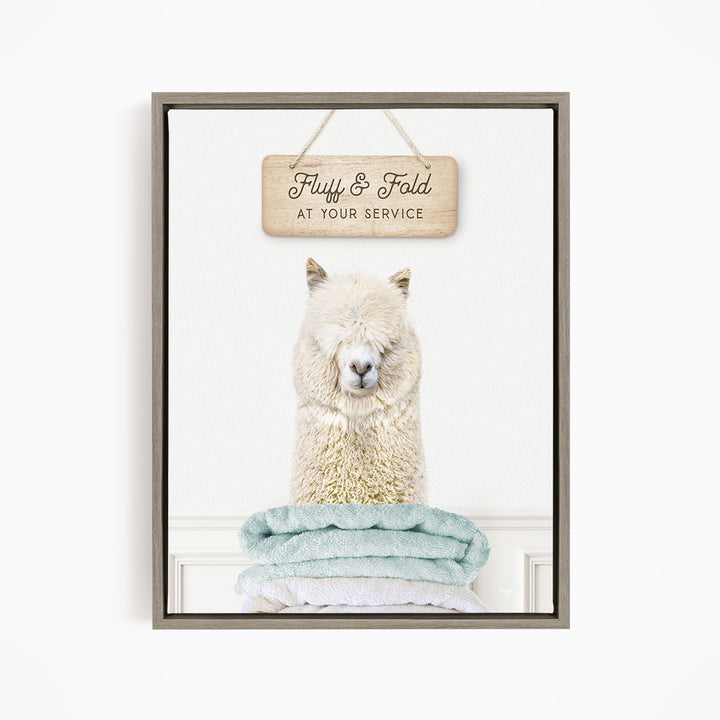 A fluffy white alpaca sitting on a stack of light blue towels, with a wooden sign above that says "Fluffy & Fold at your service".