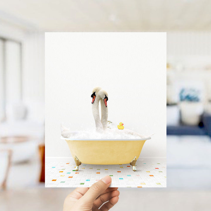 A hand holds up a framed image depicting a white swan standing in a yellow bathtub filled with bubbles, with a rubber duck floating nearby.