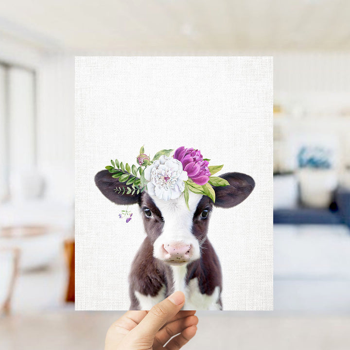 A hand is holding up a framed print of a cow with a floral crown on its head.