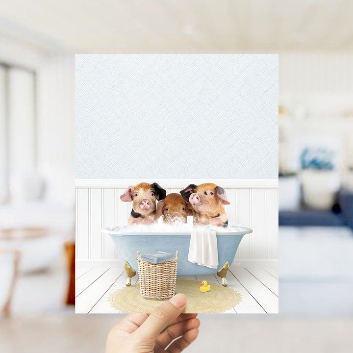 A hand holds up a framed image depicting three adorable pigs sitting in a bathtub, surrounded by a yellow rubber duck.