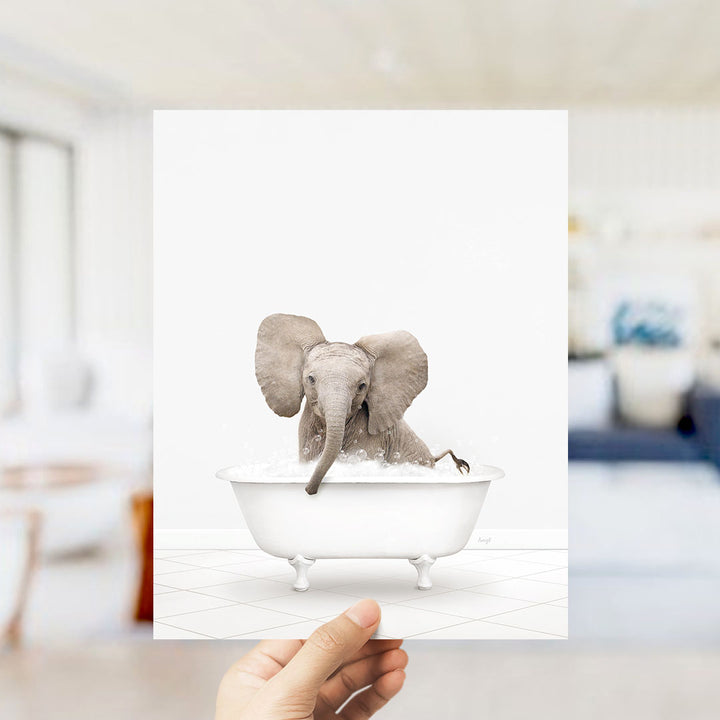 A hand holds up a framed image of a baby elephant sitting in a bathtub filled with water.