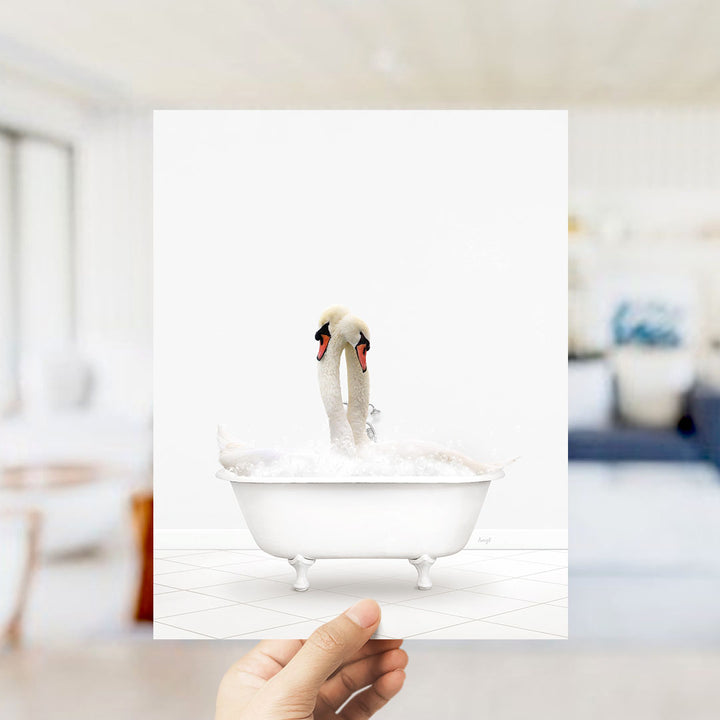 A hand holds up a framed image of two swans sitting in a bathtub filled with bubbles.