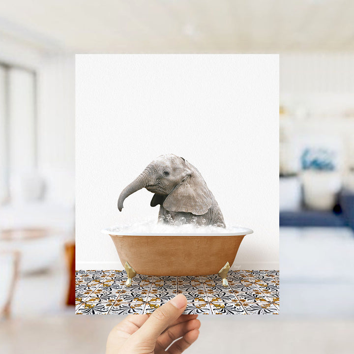 A hand holds up a framed image of a baby elephant sitting in a bathtub filled with water, surrounded by a patterned tile floor.