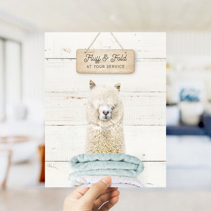 A hand holding a framed print with a picture of a fluffy white alpaca and a sign that says "Fluffy & Fold at your service".
