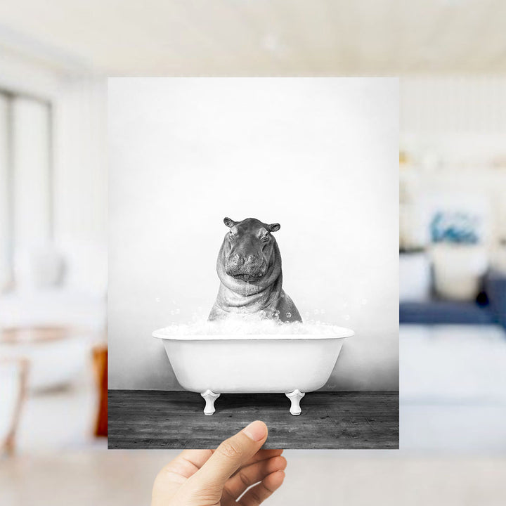 A hand holds up a black and white photograph of a hippopotamus sitting in a bathtub filled with water, creating a whimsical and playful scene.