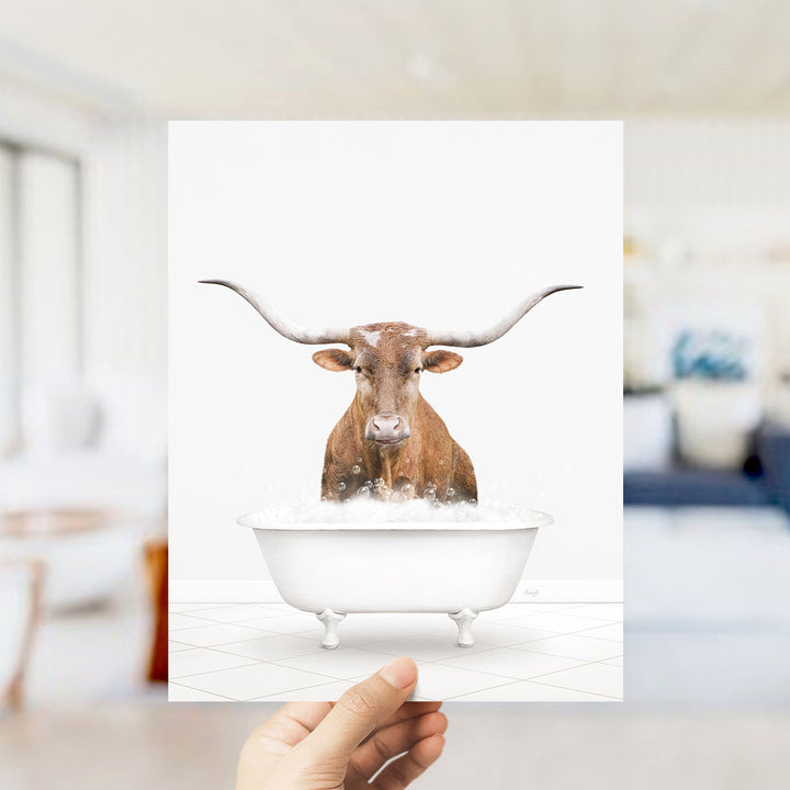 A hand is holding up a framed image of a brown cow with long horns sitting in a white bathtub filled with bubbles.
