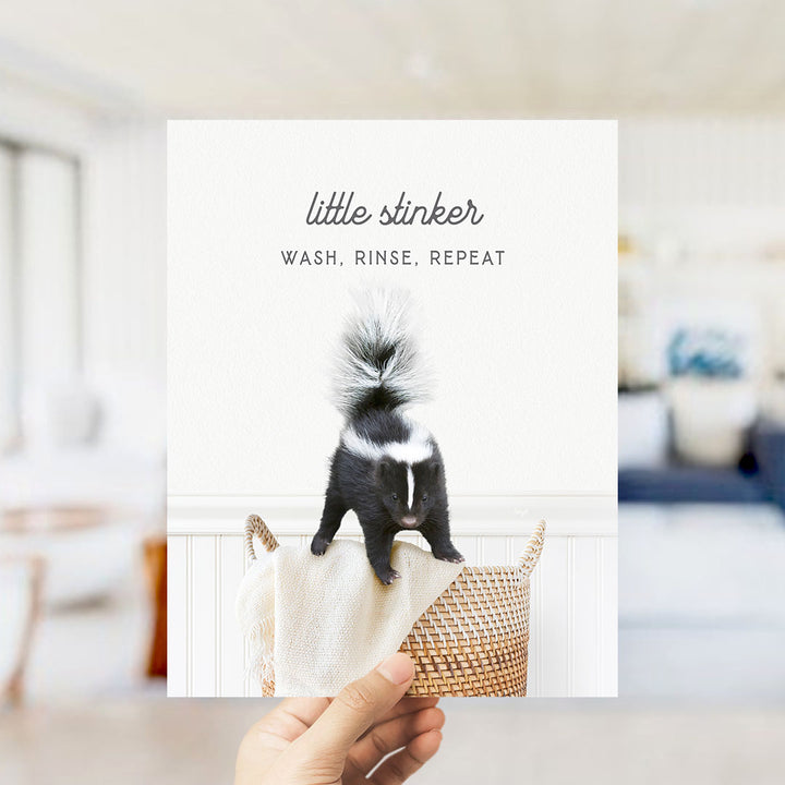 A hand holding a photo of a black and white skunk standing on a woven basket.