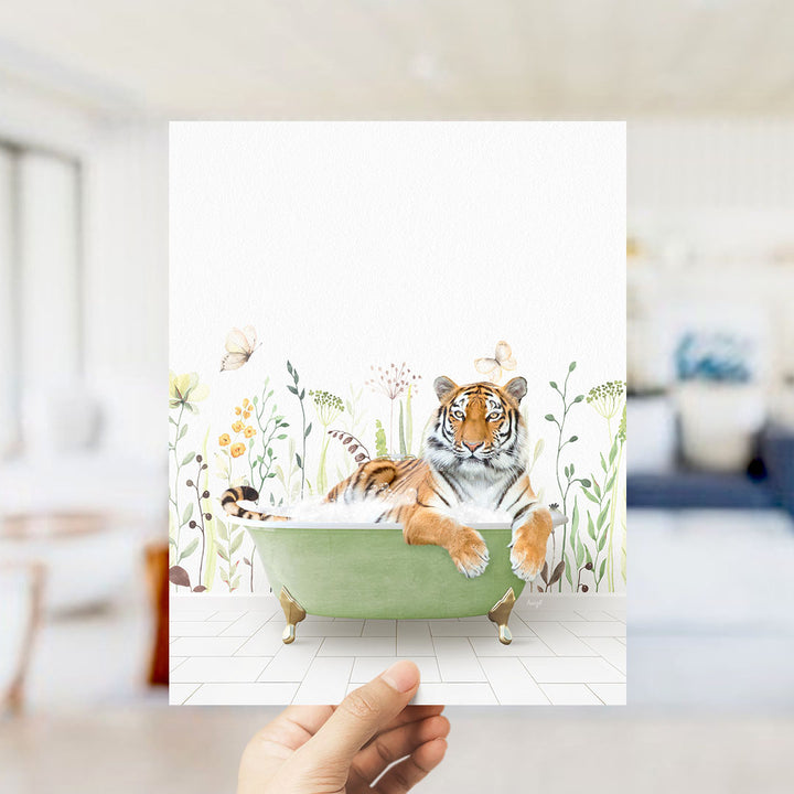 A hand holds up a framed image depicting a tiger lying in a green bathtub surrounded by flowers and butterflies.