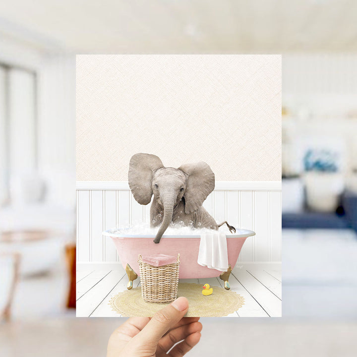 A hand holds up a framed image depicting a baby elephant sitting in a pink bathtub filled with bubbles, surrounded by a yellow rubber duck and a white towel.