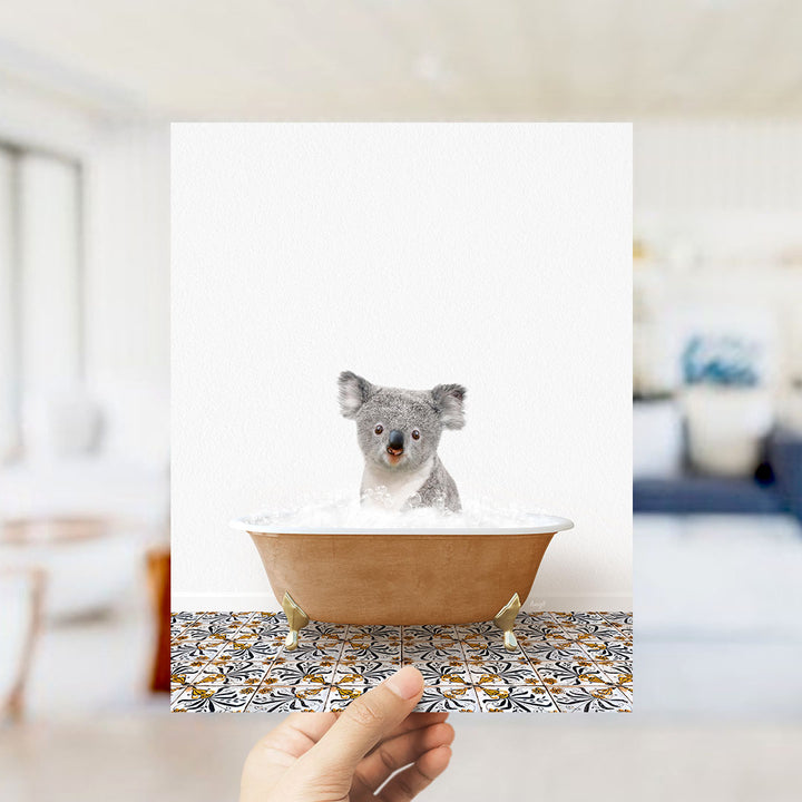A hand holds up a framed image of a koala bear sitting in a bathtub filled with bubbles, with a blurred background suggesting a bathroom setting.