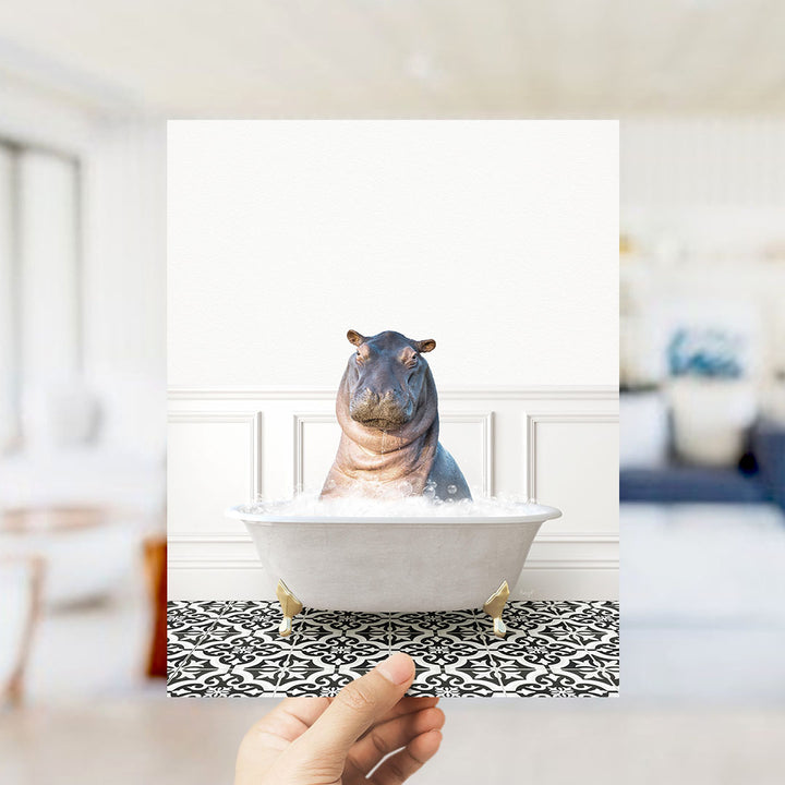 A hand holds up a framed photograph of a hippopotamus sitting in a bathtub filled with water, creating a whimsical and playful scene.