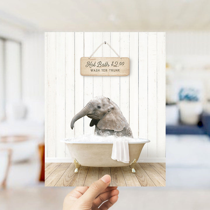 A hand holding a framed image of a baby elephant sitting in a bathtub, with a sign above it that says "Hot Bath $2.00 WASH EYE TRUNK".
