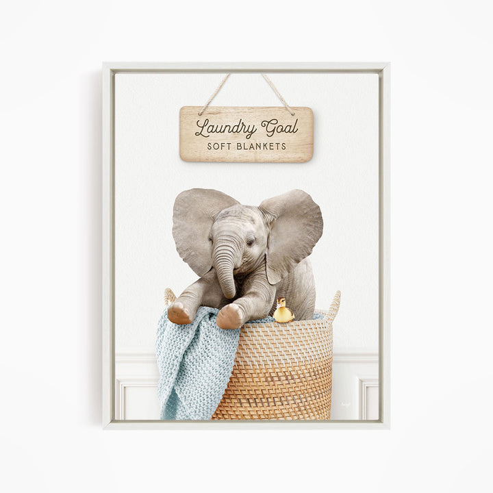 A framed image of an elephant sitting in a woven basket, with a sign above it that says "Laundry Goal Soft Blankets".