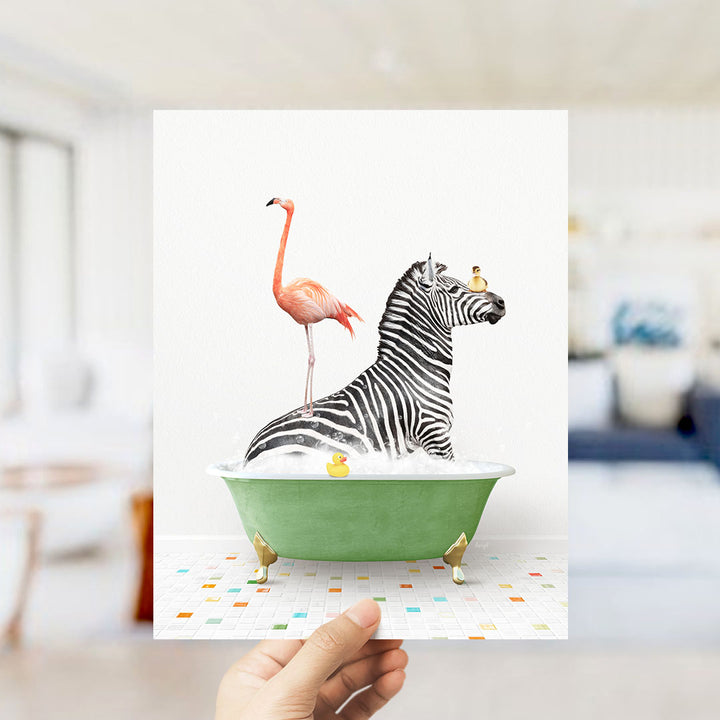 A hand holds up a framed image depicting a zebra sitting in a green bathtub, with a pink flamingo standing on its back and a yellow rubber duck floating in the water.