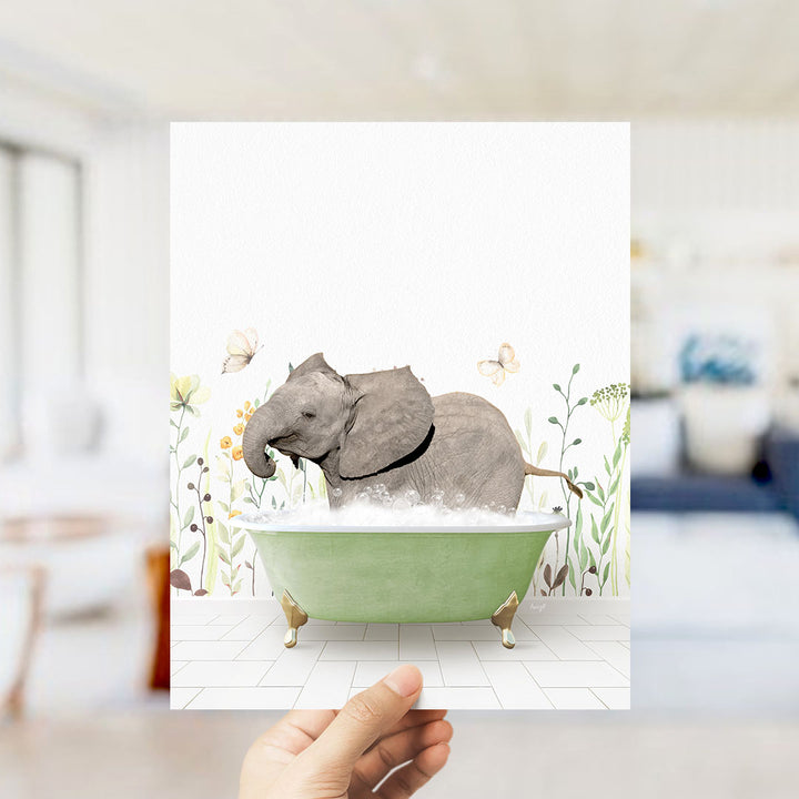A hand holds up a framed image depicting a baby elephant sitting in a green bathtub filled with bubbles, surrounded by flowers and butterflies.