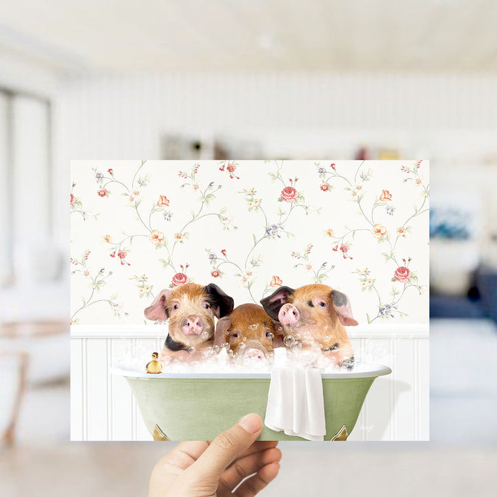 A hand holding a framed image of three adorable pigs sitting in a bathtub, surrounded by a floral pattern.