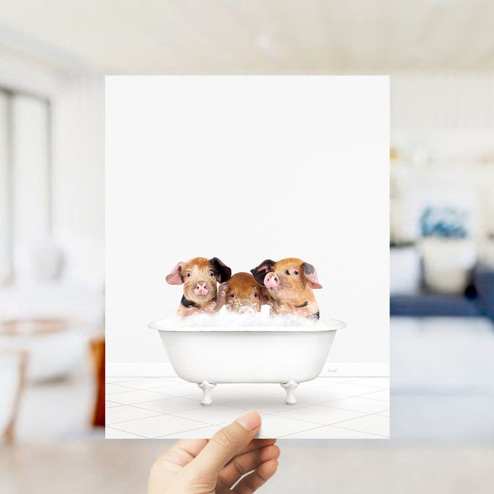 A hand holds up a framed image depicting three adorable pigs sitting in a bathtub, with a blurred background suggesting a bathroom setting.