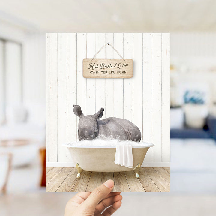 A hand holding a framed image of a rhinoceros sitting in a bathtub, with a sign above it that says "Hot Bath $2.00 WASH YER LITT L horns".
