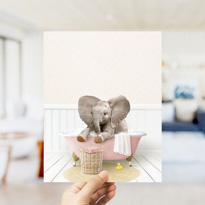 A hand holds up a framed image depicting a baby elephant sitting in a pink bathtub, surrounded by a yellow rubber duck and a white towel.