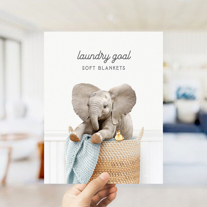 A hand holding a picture of a baby elephant sitting in a woven basket, with a blanket and a yellow duck nearby.