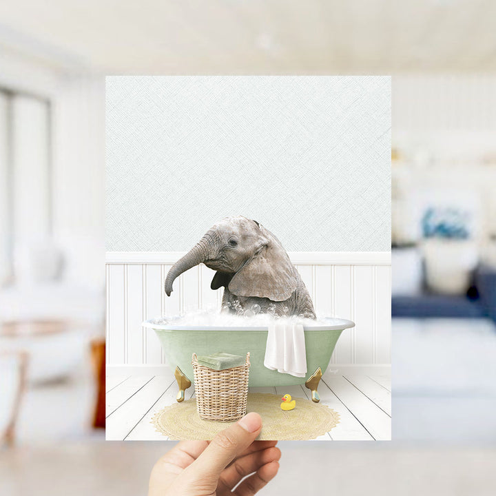 A hand holds up a framed image depicting a baby elephant sitting in a bathtub filled with water and bubbles, surrounded by a yellow rubber duck.