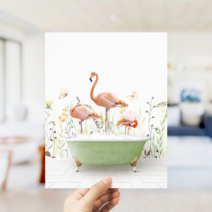 A hand holds up a framed image depicting a pink flamingo standing in a green bathtub filled with bubbles, surrounded by flowers and butterflies.