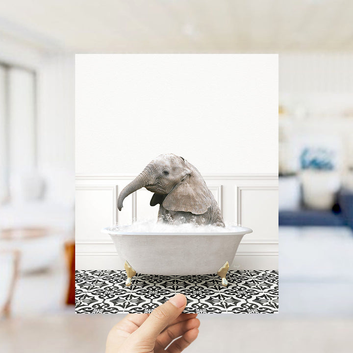 A hand holds up a framed photograph of a baby elephant sitting in a bathtub filled with water, surrounded by a black and white patterned floor.