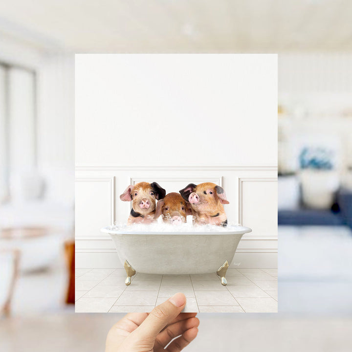 A hand holds up a framed image of three adorable pigs sitting in a bathtub, with a blurred background suggesting a bathroom setting.