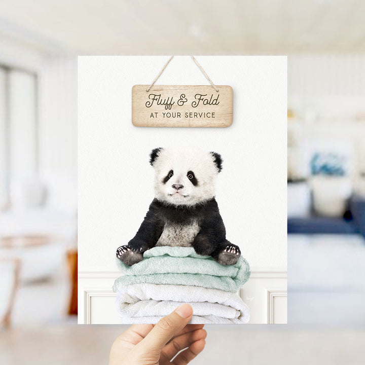 A hand holding a framed print of a panda bear sitting on a stack of towels, with a wooden sign above that says "Fluffy & Fold at your service".