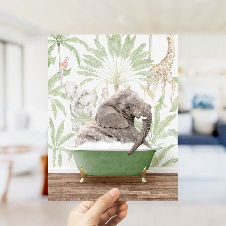 A hand holds up a framed print featuring an elephant sitting in a green bathtub, surrounded by tropical foliage and other animals.
