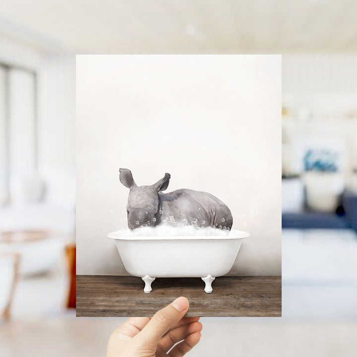 A hand holds up a framed image of a rhinoceros sitting in a bathtub filled with water, creating a whimsical and playful scene.